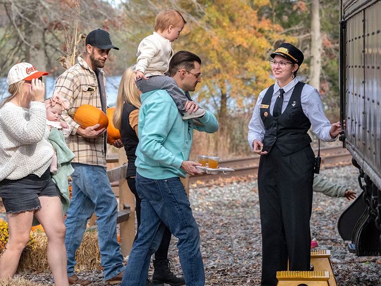 Families carrying pumpkins board a passenger train as a railroad crew member assists them during a fall outing.