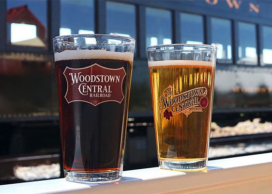 Two glasses of beer with Woodstown Central Railroad logos sit on a ledge in front of a blurred train.
